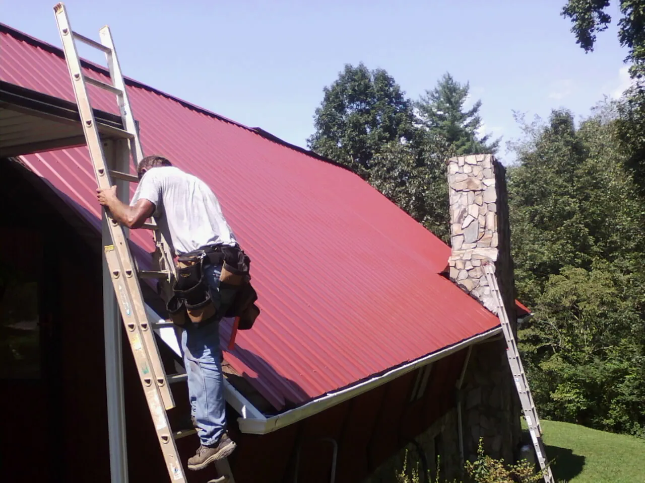 Roofer on a ladder beside a red metal roof and stone chimney — Soffit & Fascia Repair by Roofing Brothers in Watsonville